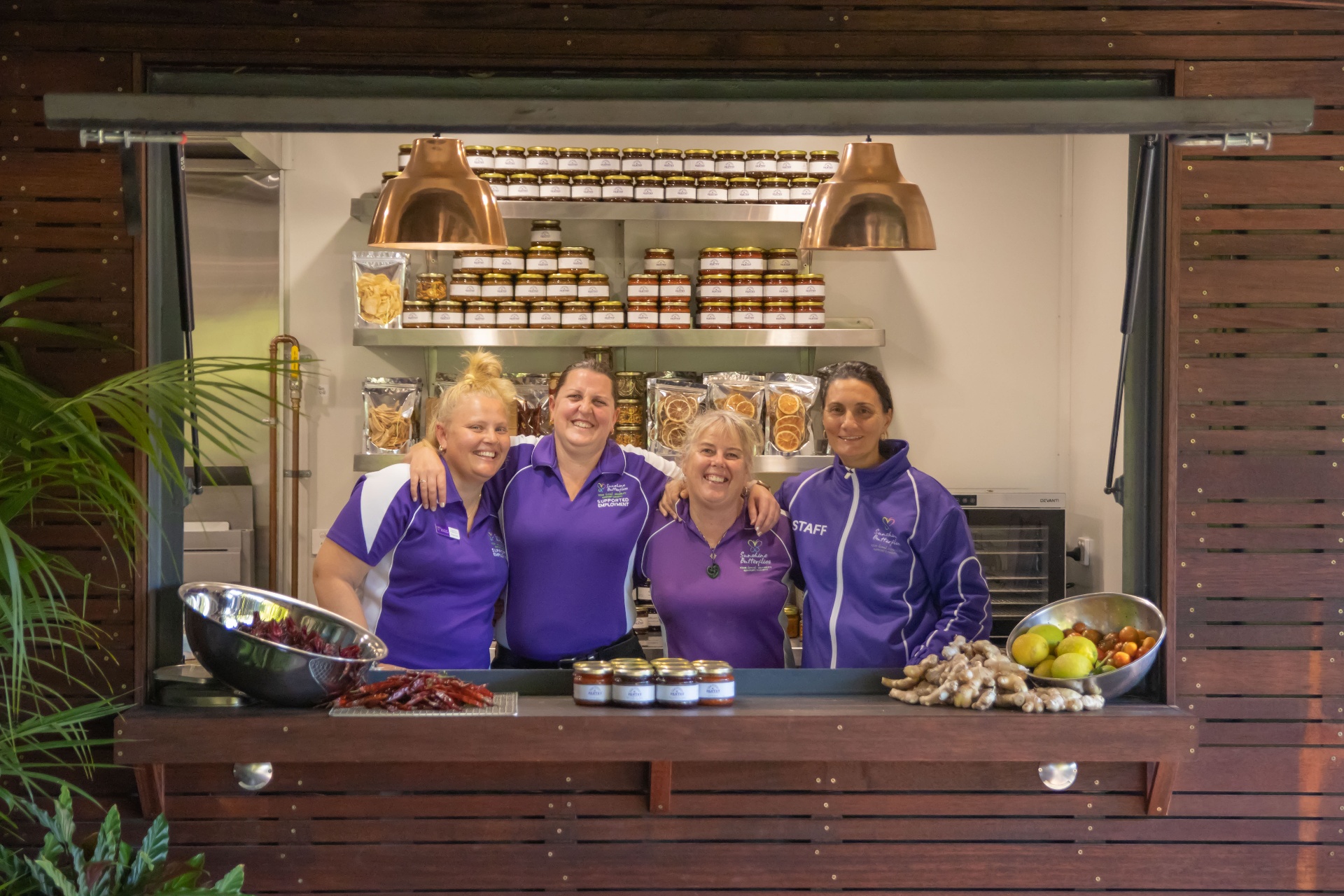 The Sunshine Butterflies Pantry team smiling behind the counter with shelves of preserves